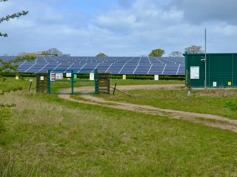 Rows of ground-mounted solar panels on a grassed site with a green substation kiosk and access track