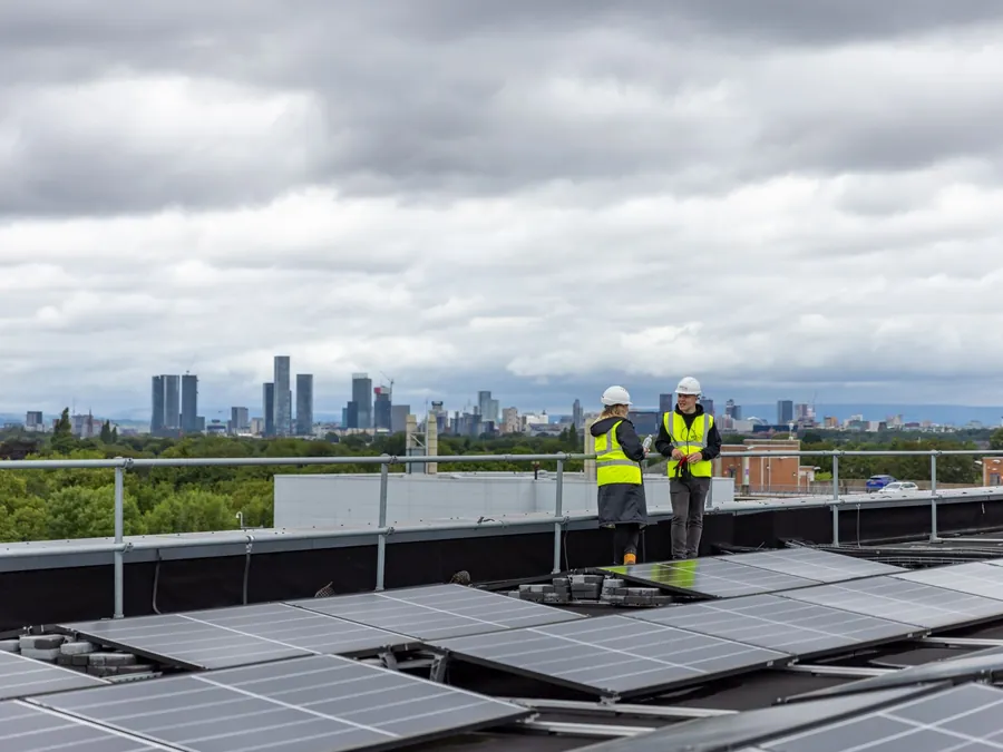 Two solar engineers in hi-vis standing on a flat roof among newly fitted panels, with a city skyline in the distance