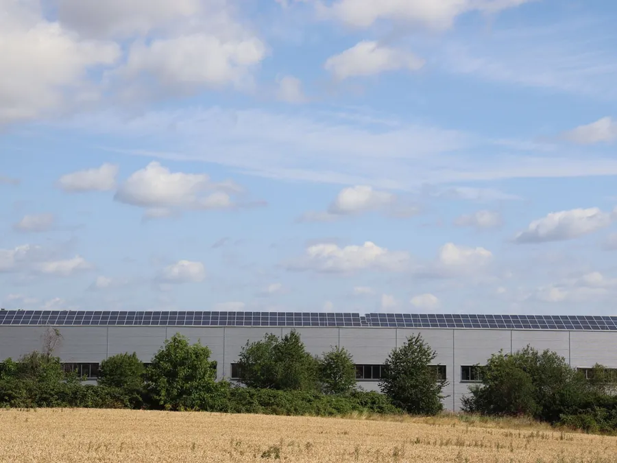 Long warehouse roof lined with solar panels, seen across a stubble field on the edge of an industrial estate